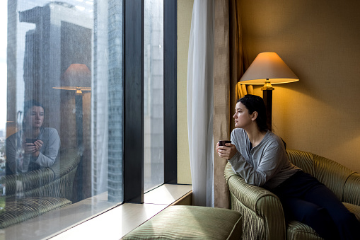 Young woman wearing pajama, holding coffee cup and looking at cityscape through the window in luxury penthouse apartment or hotel room