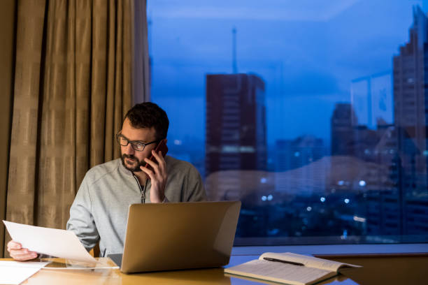 istock Handsome man using laptop and mobile phone in penthouse or luxury hotel room doing paper work report. Businessman in skyscraper apartment against night city background working late at nigh. 1759763236