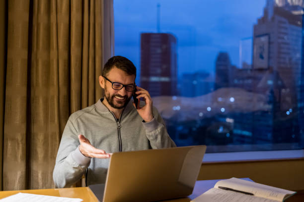istock Handsome man using laptop and mobile phone in penthouse or luxury hotel room doing paper work report. Businessman in skyscraper apartment against night city background working late at nigh. 1759763240