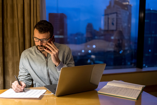 Young handsome man using laptop and mobile phone in penthouse or luxury hotel room doing paper work report. Businessman in skyscraper apartment against night city background working late at nigh.