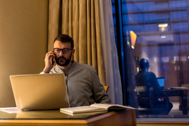 istock Handsome man using laptop and mobile phone in penthouse or luxury hotel room doing paper work report. Businessman in skyscraper apartment against night city background working late at nigh. 1759763324