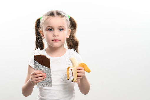 A child is offered chocolate and chooses a banana, on a white background a man's hand gives a chocolate to a little girl holding a banana in her hand, copy space and white background.