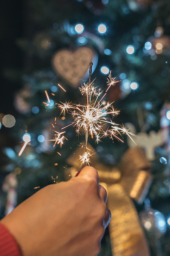 Woman holding a sparkler to celebrate Christmas. Unrecognizable young girl raising a lighted sparkler making a wish on New Year's Eve while looking at the Christmas tree.