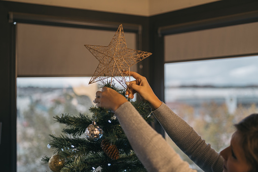 Young woman placing a shiny decorative star on the top of the Christmas tree.