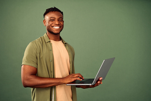Friendly african american guy standing over green studio background, smiling at camera and holding wireless laptop. Happy male person using modern computer for remote work.