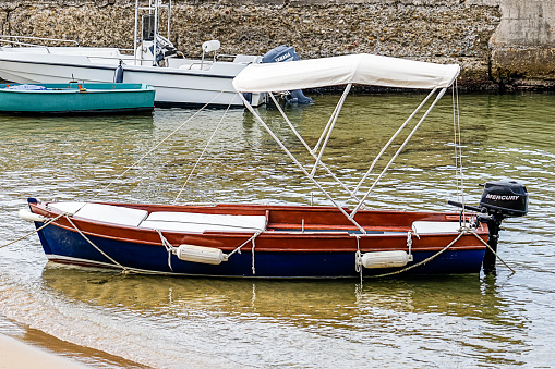 Bow of small blue row boat anchored in the calm bay. White trim around the top. Blue base with a rust colored bottom which reflects in the calm water. Pier pilons out of focus in the background