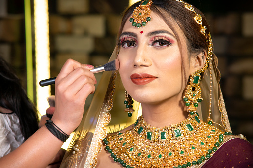 Close-up portrait of beautiful Indian bride getting ready for her wedding day at the parlor by makeup artist. She is looking at the camera with smile.