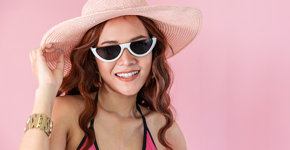 A vibrant young Asian woman epitomizes summer beauty. Her sun-kissed skin, paired with chic sunglasses and a sunhat, captures the spirit of a young adult reveling in the warmth of summer