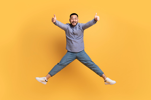 Joyful Young Asian Man Jumping In Air And Showing Thumbs Up At Camera, Positive Millennial Guy Recommending Something And Having Fun Over Yellow Studio Background, Full-Length, Copy Space