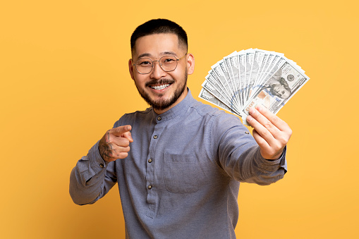 Happy Asian Man Holding And Pointing At Dollar Cash Fan In His Hand, Cheerful Male Showing Money Banknotes While Standing Over Yellow Studio Background, Celebrating Financial Success And Profit