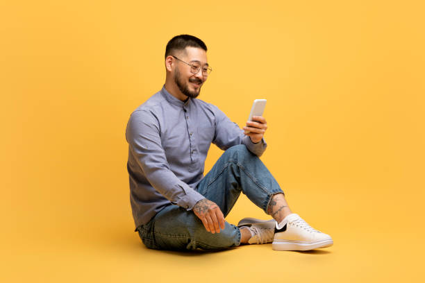 istock Smiling Asian Man Using Smartphone While Sitting On Floor Over Yellow Background 1759880029