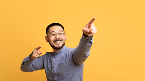 Gotcha. Cheerful asian man pointing away with two hands, handsome millennial male in eyeglasses showing direction with fingers, indicating copy space on yellow studio background, panorama