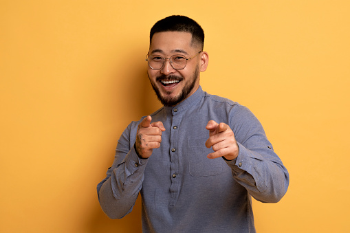 Gotcha. Cheerful Handsome Asian Man Poiting At Camera With Two Hands, Positive Millennial Guy In Eyeglasses Indicating Somebody And Smiling, Having Fun On Yellow Studio Background, Copy Space