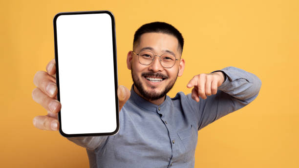 istock Mobile Ad. Smiling Young Asian Man Pointing At Blank Smartphone In Hand 1759880675