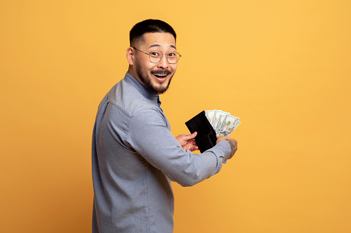 Money Concept. Happy asian man holding open wallet with cash and turning at camera, standing on yellow studio background, smiling milllennial guy got big profit, celebrating successful investment