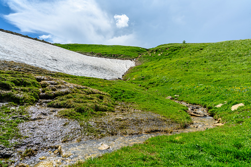 Mountain stream with a waterfall in the Alpine mountains. Beautiful landscape with mountains, green grassy meadows and a hiking trail in springtime.