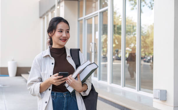 istock Attractive young Asian female student stands holding a book and using her phone to make an appointment with a college friend. 1759903976