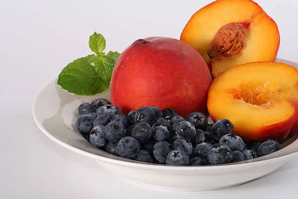 fresh whole and halved peaches served in a white bowl with a prig of mint and a healthy dose of blueberries