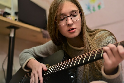 A teenage girl with glasses emotionally plays the guitar and sings songs. Creative recreation at home. White spots on the teeth.