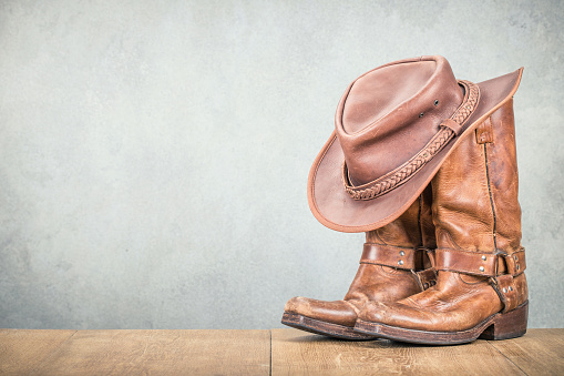 Vintage cowboy hat and old leather Wild West boots front concrete wall background. Retro style filtered photography