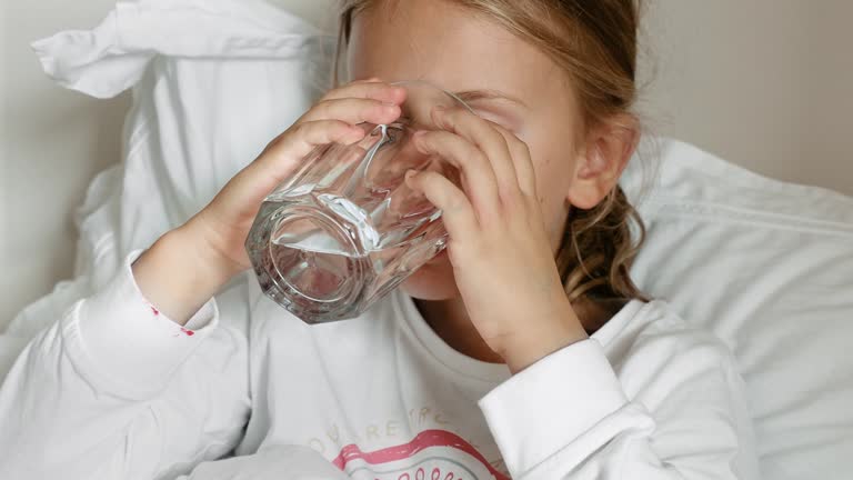 Health stay hydrated concept. Thirsty Child Drinking Water at home in pajamas. Kid drink fresh pure water glass cup siting on bed. Take care body. Morning routing. Hydration tips for children