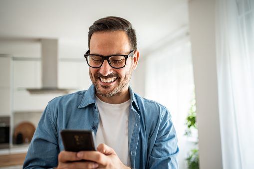 Happy man using mobile phone while in his apartment, surfing internet or sending sms message