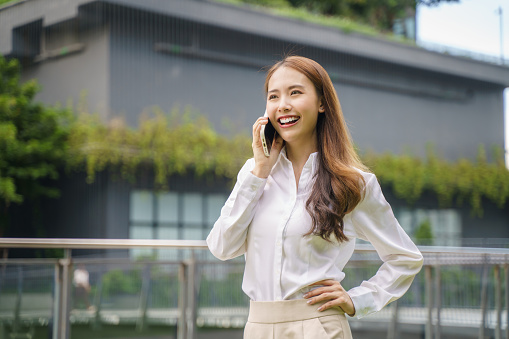 Young beautiful Asian business woman using smartphone talking with friend or colleague outdoor urban background.