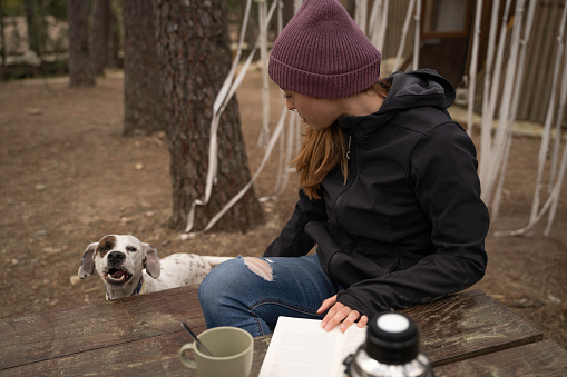 Outside a Glamping , The girl is with her dog and drinks coffee while reading a book on a wooden table outdoors during the fall.