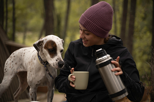 Girl in winter hat is sitting while drinking coffee with her dog outdoors.
