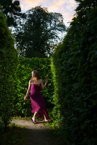 Young woman with a long blond hair running through the hedge maze late in the evening.