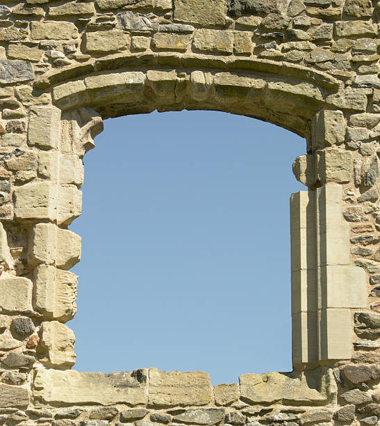 A partially repaired window in a a wall of a ruined 13th-century priory framing a blue sky.