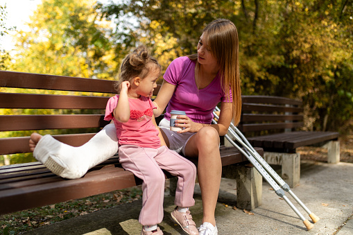 A mid adult woman with a cast on her leg sitting on a bench looking at her daughter in park. Girl is sitting next to her mother