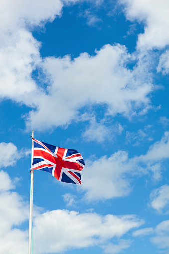 vertical colour image of the typically British union Jack flag blowing in the breeze on a glorious summer's day against a blue sky with white fluffy clouds. Flag seen on white flag pole. Lots of copy space.