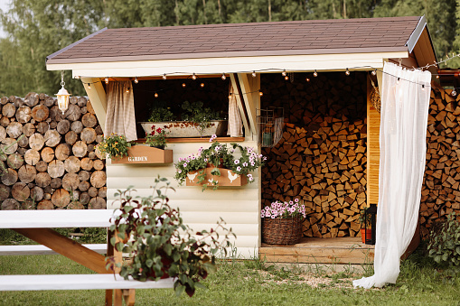 A cozy wooden shed with a lot of firewood in a tidy green yard with potted plants and flowers. Concept for home improvement, gardening, or rural lifestyle. Copy space on the left.