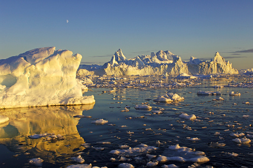 The golden light of the midnight sun illuminates Icebergs near Ilulissat off the coast of Greenland. The Moon shines above