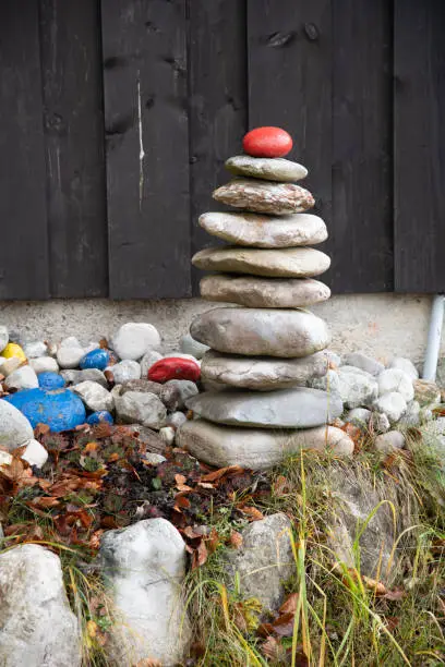 A small stone tower or cairn erected in a garden with a red stone at the top in front of a brown wooden wall. In the foreground you can see a little grass.