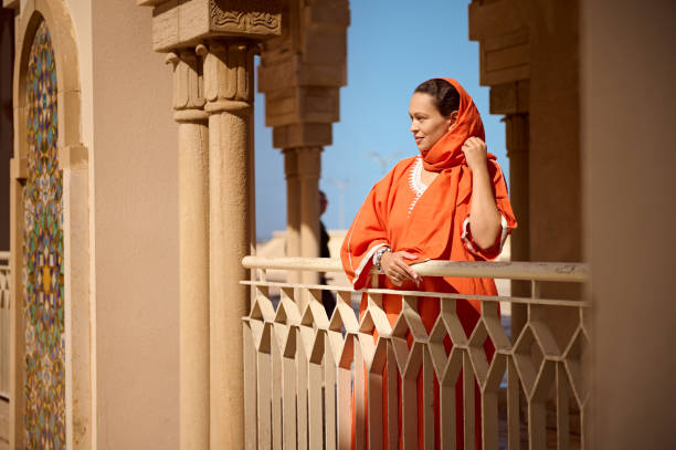 Beautiful woman in stylish oriental attire and covered head in scarf of orange color, looking into distance, in a mosque