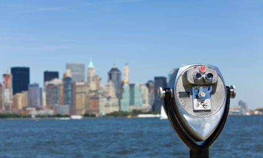 A tourist  telescope on the  Ellis Island and New York City Skyline with the Hudson River and the  Lower Manhattan skyscrapers in the background on a sunny summer day.