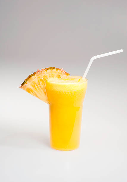 A glass with freshly squeezed pineapple juice. The glass is decorated with a slice of pineapple and a white straw. Background is a gradient from grey to white.