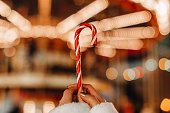Female hands holding red white cane sweet lollipop against the background of golden garland lights.