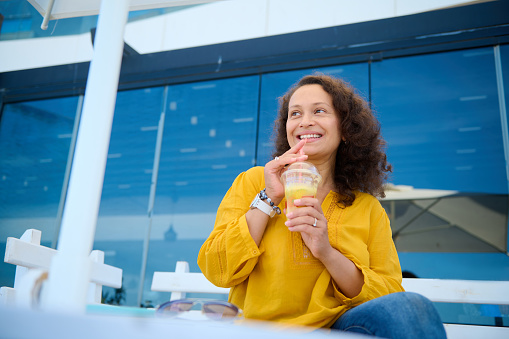 View from the bottom of happy multi-ethnic woman, taking a sip of freshly squeezed healthy fruit smoothie, smiling broadly looking aside, sitting in an outdoor terrace against glass windows background