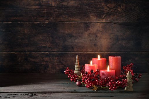 Third advent wreath with red candles, three are lit, decoration with berries, Christmas balls and small wooden trees, dark rustic background, copy space, selected focus, narrow depth of field