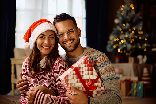 Young happy couple with a gift box during Christmas day at home looking at camera.