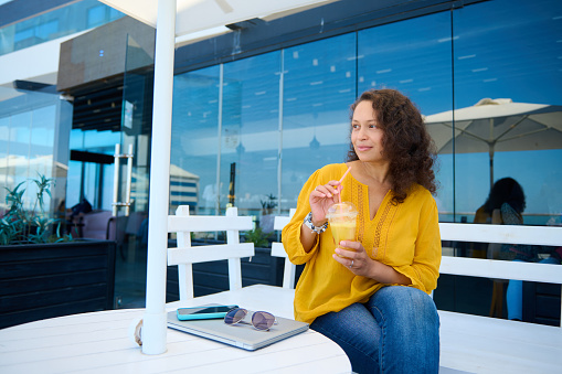 Attractive curly haired multi-ethnic middle aged woman holding glass of freshly squeezed orange juice, looking dreamily aside, sitting on a white wooden bench with mobile phone and laptop on the table