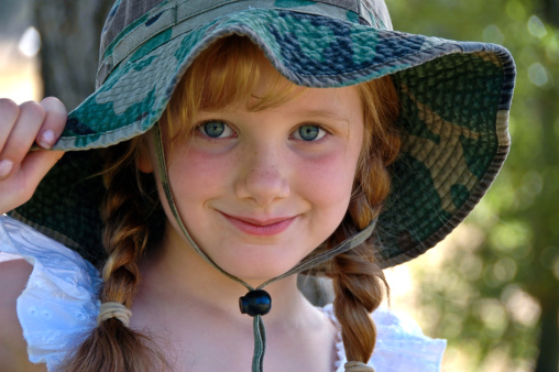 Head shot of an adorable seven year old girl with red hair in braids wearing a camouflage hat. Click photo below to see more of this model.