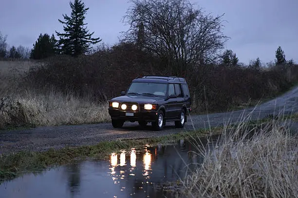 A Landrover Discovery with its fog lights on driving on a gravel road next to a pond