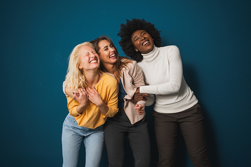 Three multicultural female friends smiling and embracing each other in a studio. Group of vibrant young women enjoying themselves while standing against a blue background.