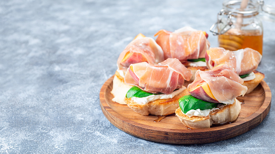 Prosciutto peach bruschetta on wooden plate. Crostini with cream cheese, basil leaf, peach wrapped in prosciutto, topped with honey, horizontal, copy space