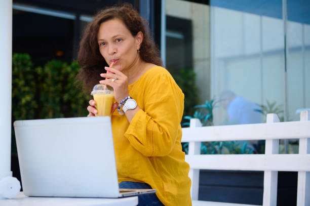 istock Attractive middle aged woman drinking a freshly squeezed orange juice from a straw, working remotely on laptop 1760312951