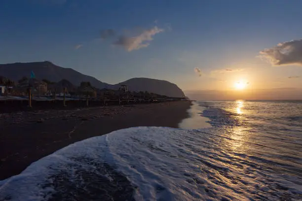 Photo of Santorini - Black beach of Perivolos with a beautiful long wave of the sea, illuminated by the first rays of the sun at sunrise. On the beach white sunbeds with umbrellas and in the background a high